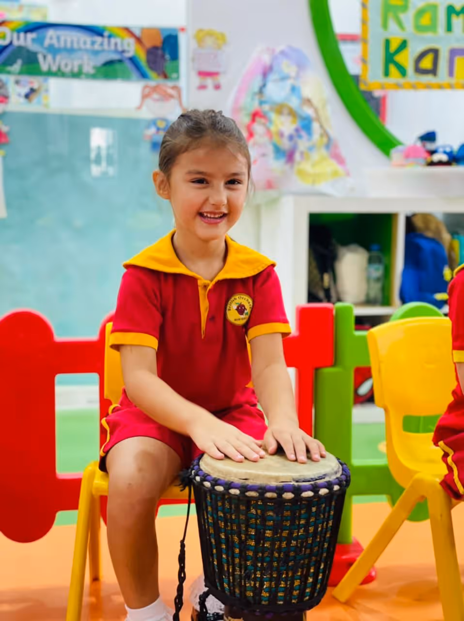 Smiling young girl in a red and yellow school uniform playing a drum while seated on a yellow chair in a colorful classroom.