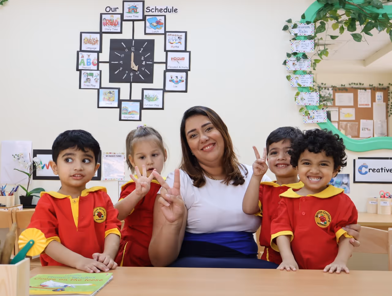 Teacher sitting with four smiling children in red uniforms at a classroom table with a schedule clock on the wall behind them.