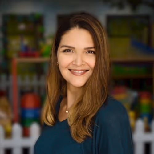 Smiling woman with long brown hair wearing a dark teal top, standing in front of a blurred colorful background.