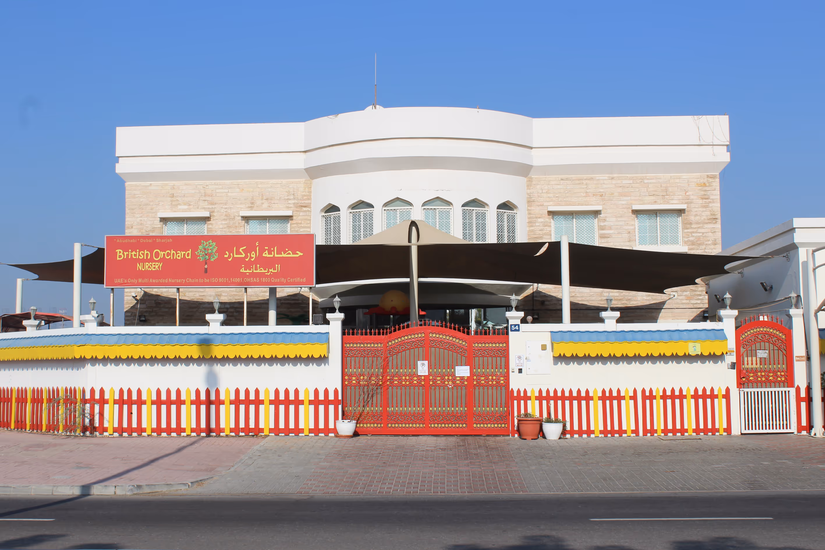 Front view of British Orchard Nursery building with red gates and a colorful fence under a clear blue sky.