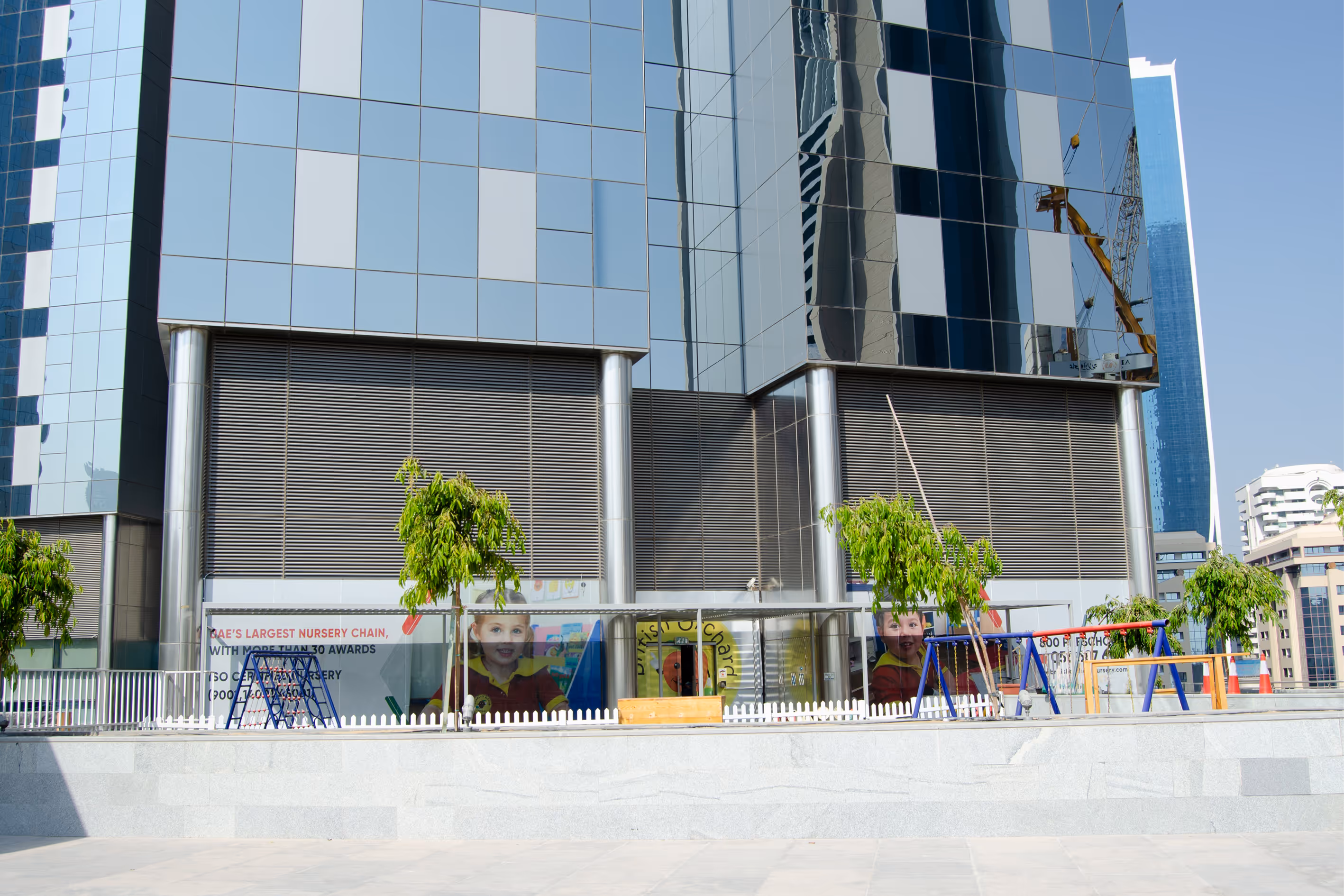 Modern glass building with a fenced outdoor play area featuring swings and small trees in front of a nursery.