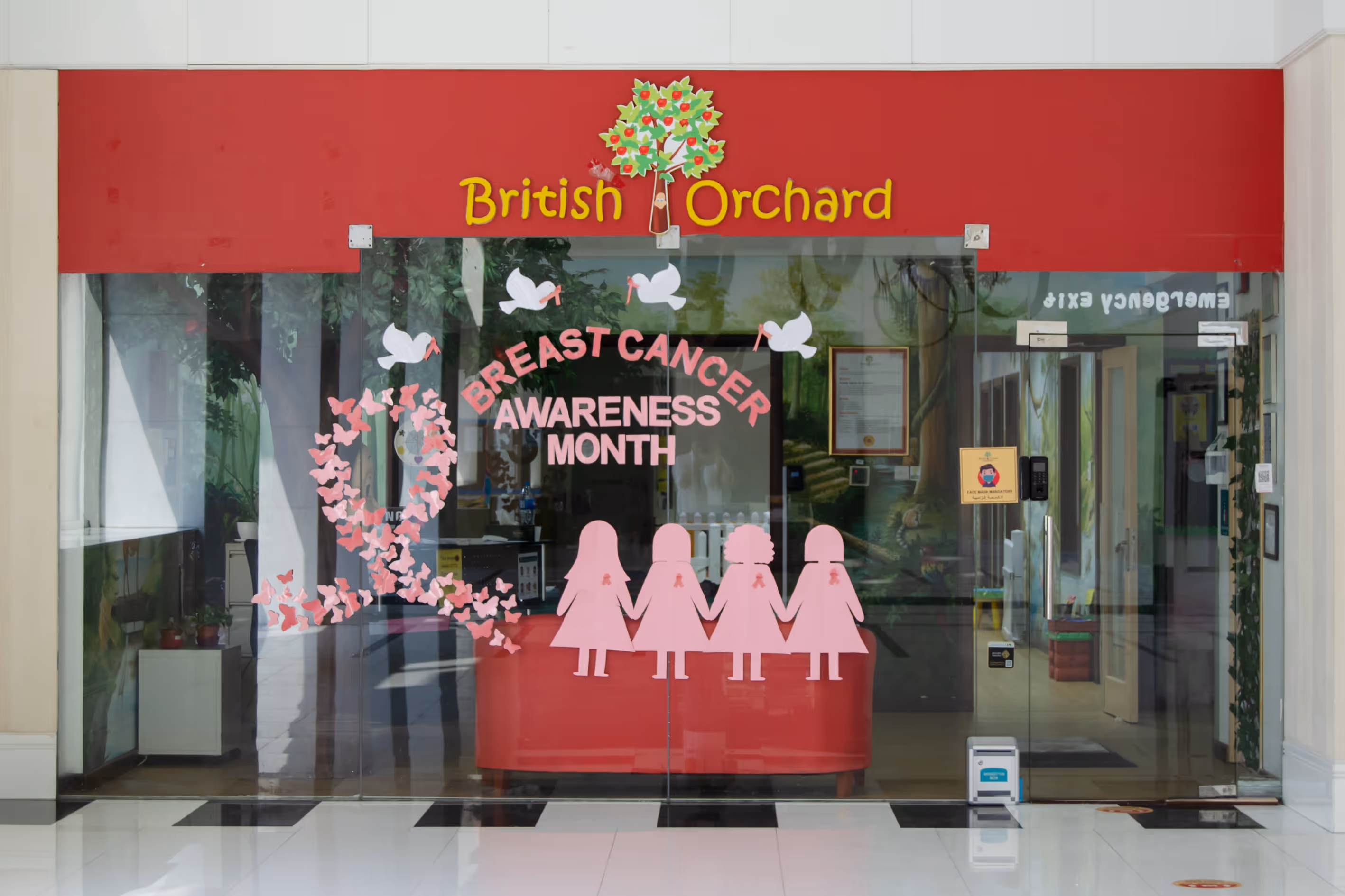 Glass entrance decorated with pink ribbons, butterflies, and silhouettes of four women holding hands for Breast Cancer Awareness Month at British Orchard Nursery.