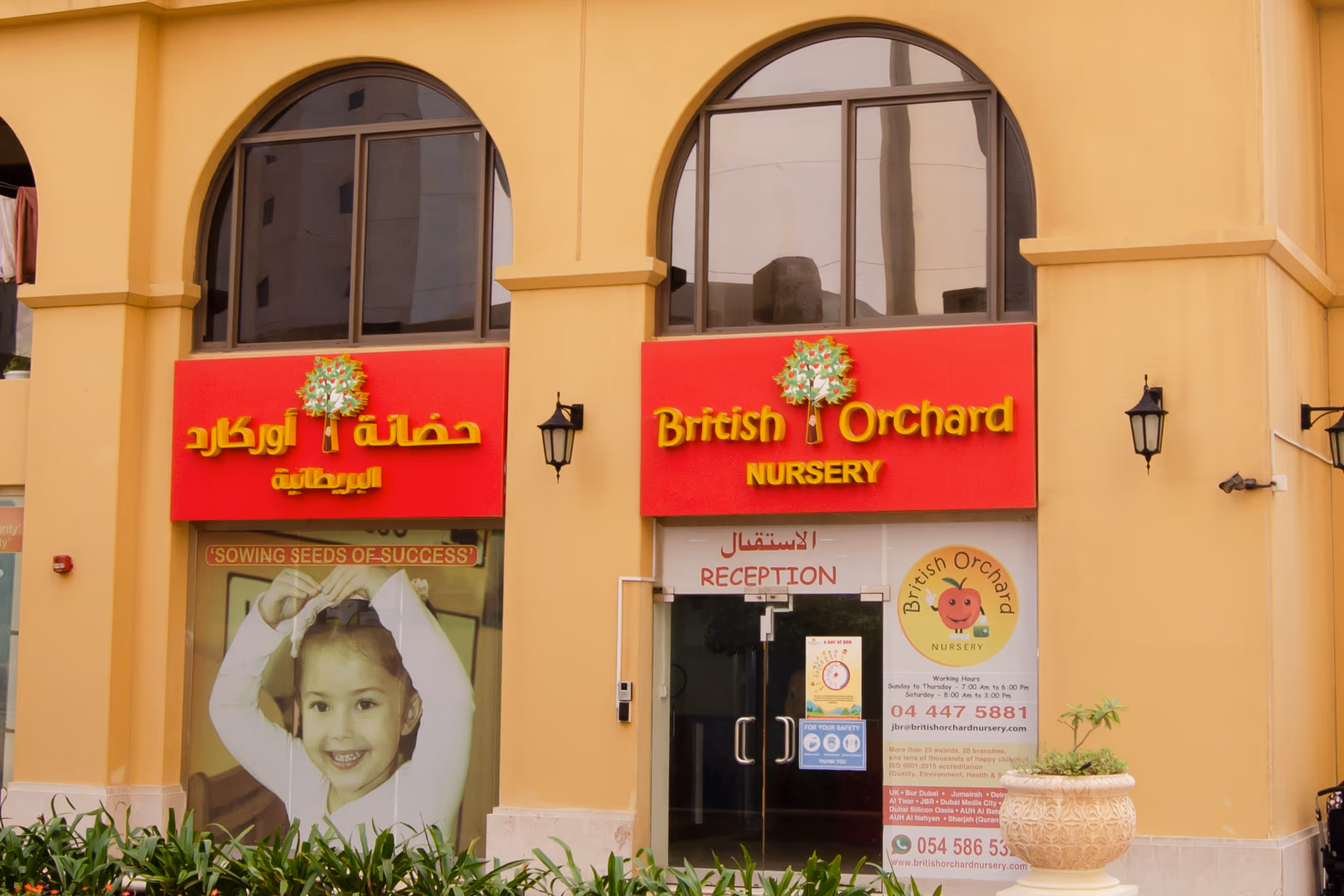 Entrance of British Orchard Nursery with red signs in English and Arabic, a smiling child poster, and a reception door.