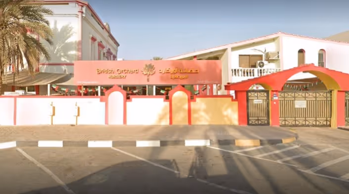 Exterior view of British Orchard Nursery with white and red walls, palm trees, and a gated entrance under a red canopy.