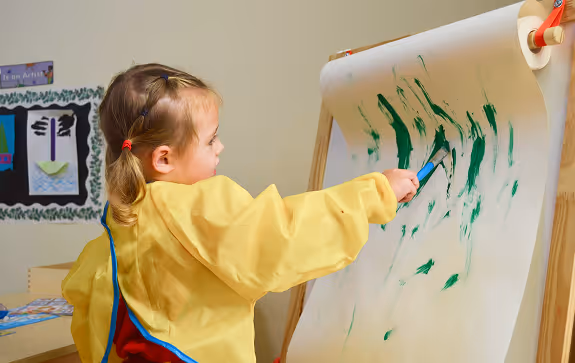 Young girl in a yellow smock painting green strokes on large paper attached to an easel indoors.