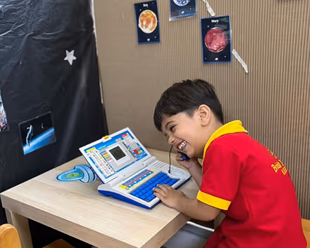 Young boy in a red shirt smiling while playing with a toy laptop and phone at a wooden desk with space-themed decorations on the wall.