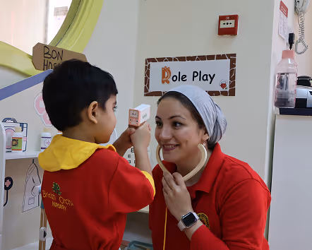 Child in red and yellow uniform playing doctor using a toy stethoscope with a smiling adult woman in a headscarf under a Role Play sign.