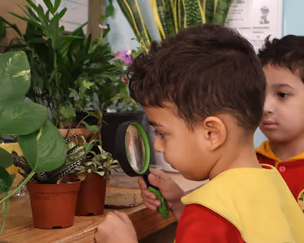 Young boy closely examining potted plants with a magnifying glass while another child watches.