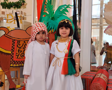 Two children wearing traditional Emirati clothing, a boy in a white kandura and red-checkered headscarf, and a girl in a white dress with red, green, and black accents, standing indoors near decorations including a camel illustration and palm tree cutout.