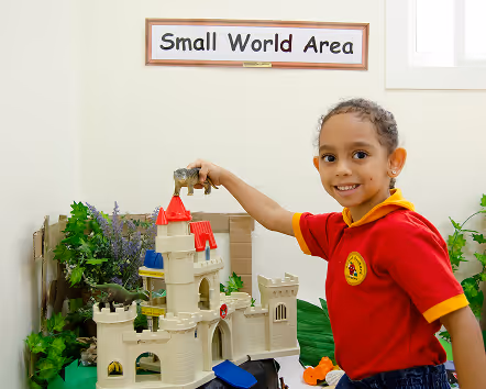 Smiling child playing with animal toys on a castle model in a play area labeled Small World Area.