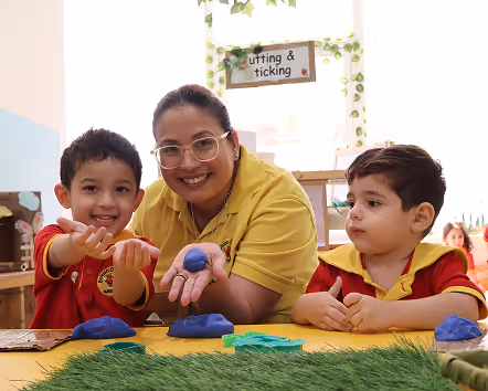 Teacher smiling with two young boys playing with blue playdough at a table in a classroom.