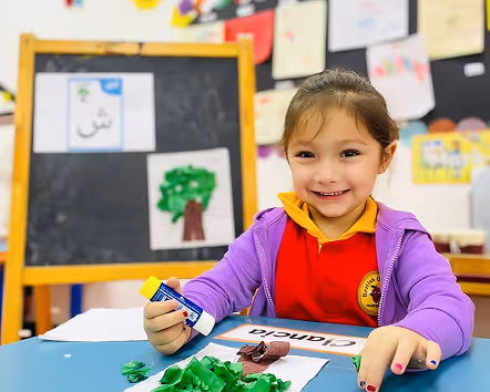 Young girl smiling while crafting with glue and green paper at a classroom table.
