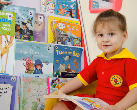 Young child in red and yellow outfit sitting and reading a colorful picture book with a display of children's books behind.