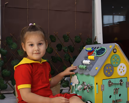 Young girl playing with a colorful sensory activity toy house featuring gears, locks, and beads.