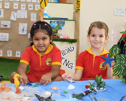 Two young girls in red and yellow school uniforms sitting at a table with sea animal toys and shells, smiling at the camera.