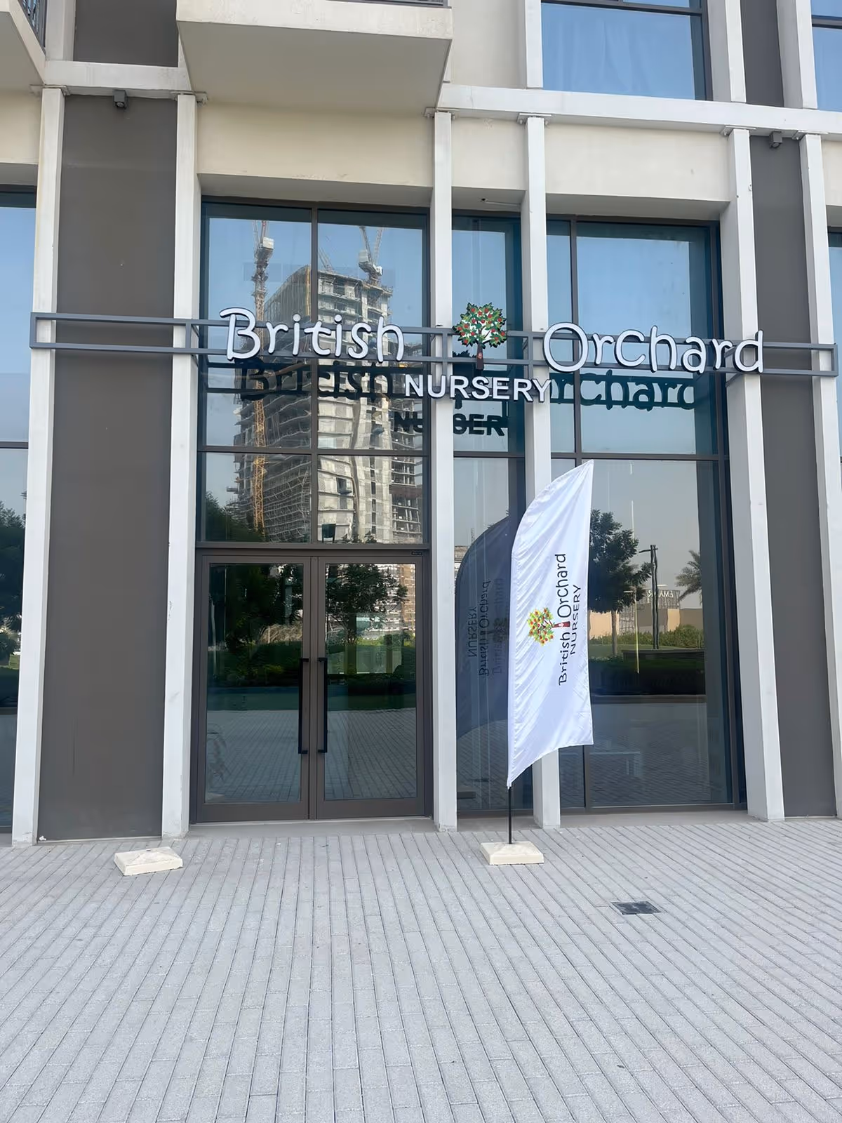 Entrance of British Orchard Nursery with glass doors, reflected building, and a flag displaying the nursery's name and logo.