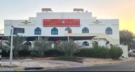 Two-story white building with blue arched windows and a red sign reading 'British Orchard Nursery' in front of a street corner with palm trees and street sign 'Al Bateen St'.