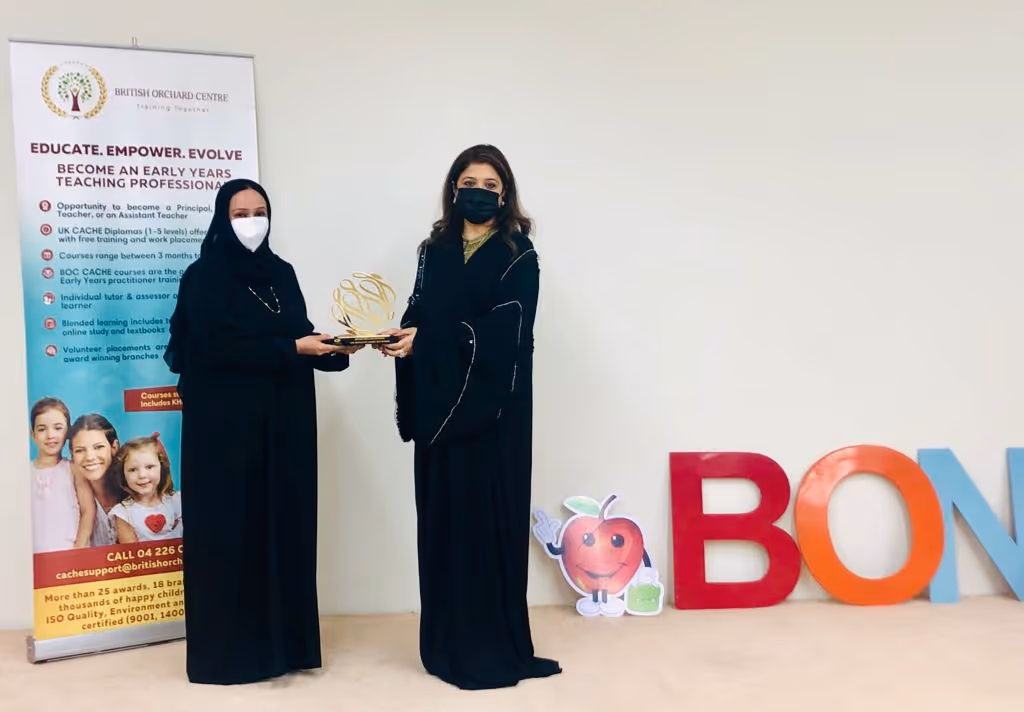 Two women in black abayas and face masks standing indoors holding a trophy, next to a banner for British Orchard Centre and colorful large letters spelling B O N.