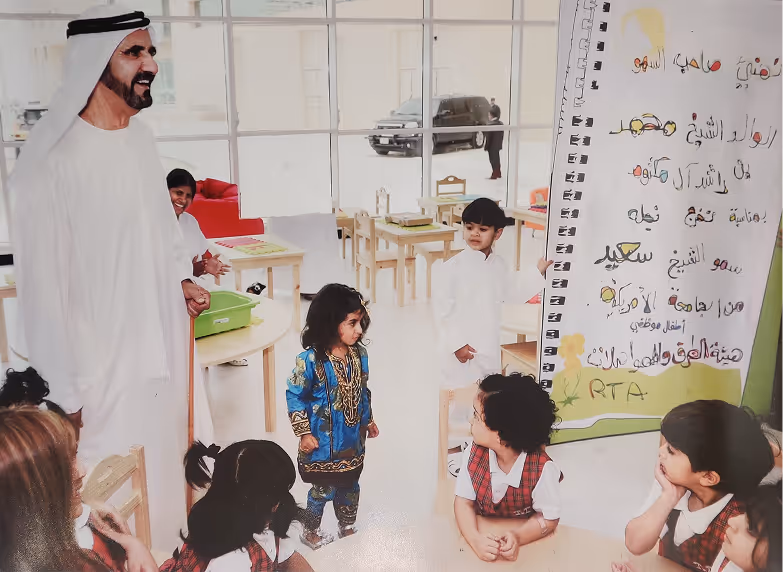 A man in traditional Emirati attire smiling and interacting with children in a classroom setting with Arabic writing on a large poster in the background.
