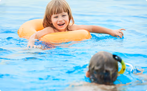 Young girl smiling and floating in a swimming pool with an orange swim ring, while a person wearing a snorkel mask is in the foreground.