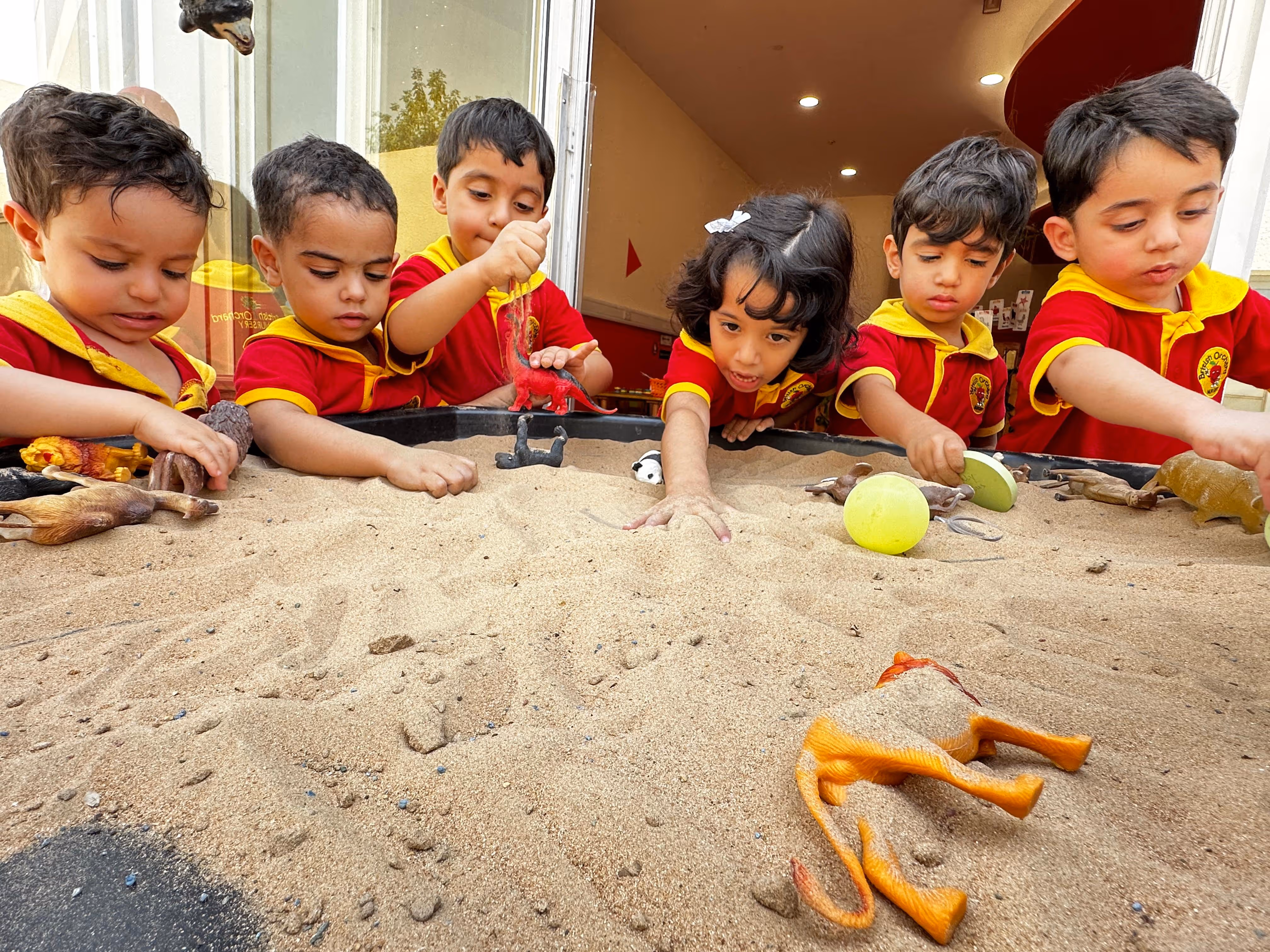 Six children in red and yellow uniforms playing with toy animals in a sand table.