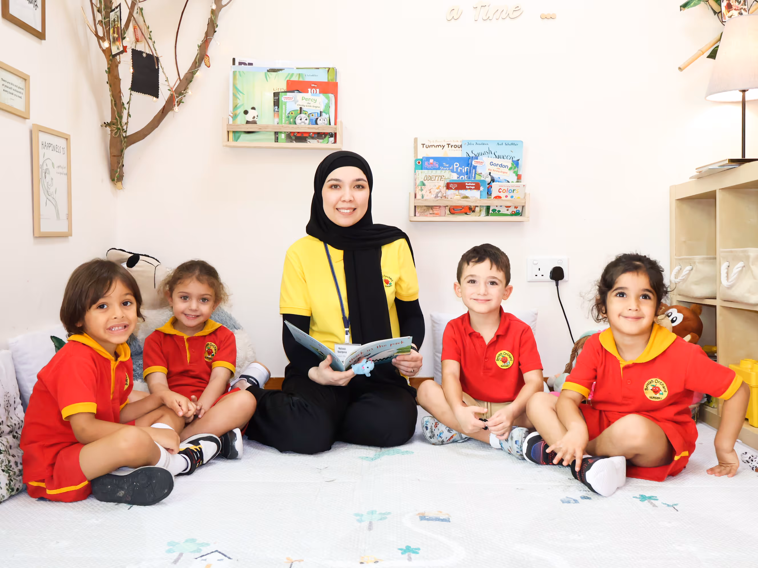 Smiling female teacher in a yellow shirt and hijab reads a book seated on the floor with four young children in red uniforms in a bright classroom.