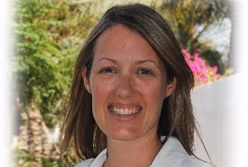 Smiling woman with long brown hair standing outdoors with greenery and flowers in the background.