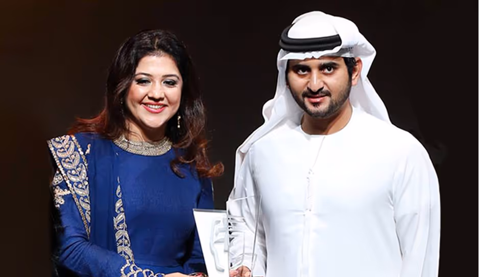 Smiling woman in blue traditional dress and a man in white kandura holding an award together.