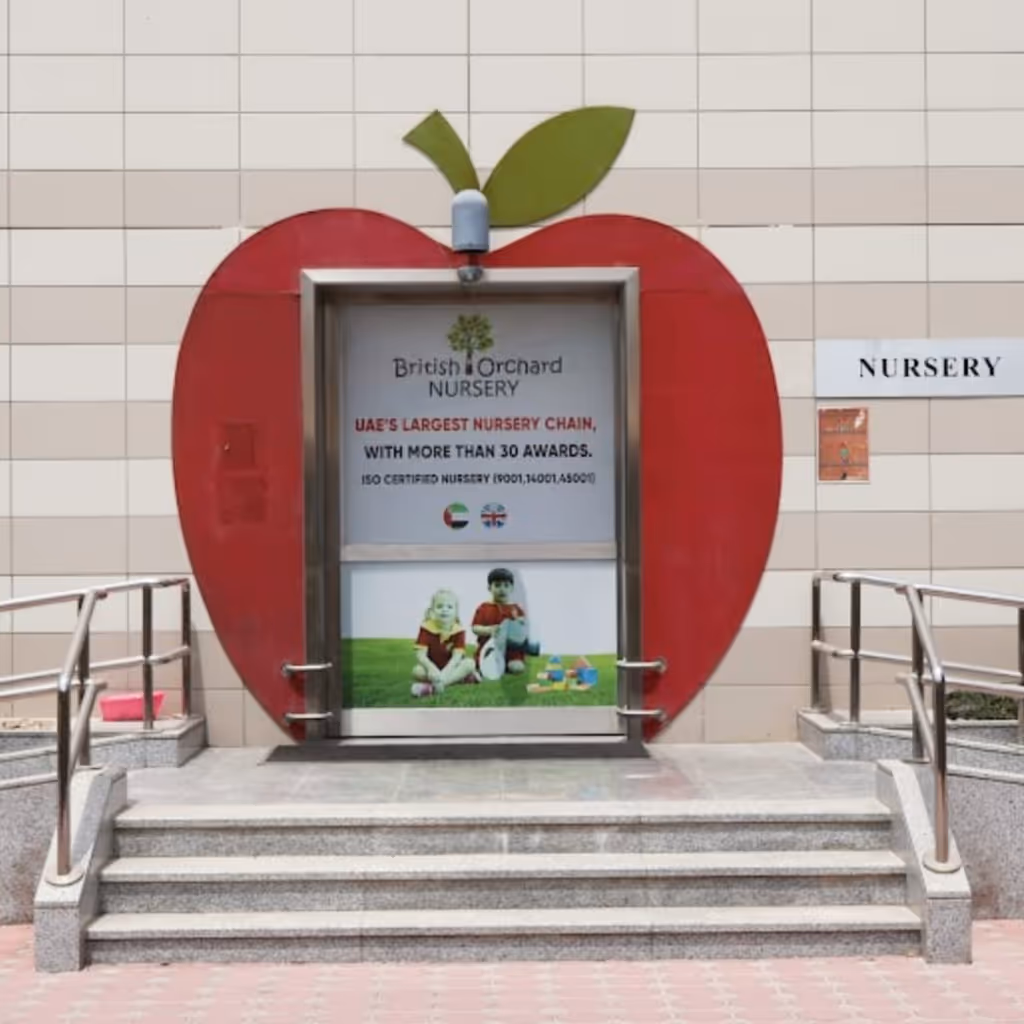 Entrance door of British Orchard Nursery framed by a red apple shape with green leaf, featuring text about UAE's largest nursery chain and children playing on grass.