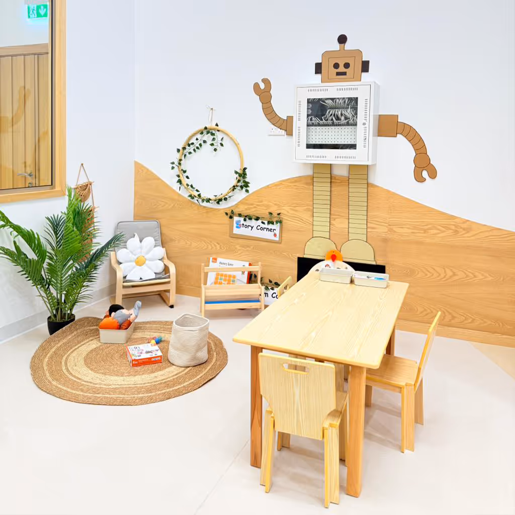 Children's reading corner with wooden table and chairs, a small armchair with a white flower cushion, a woven rug, plants, and a robot-shaped book display on the wall.