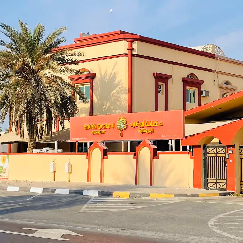 Building with beige walls and red trim with a sign that reads British Orchard Nursery in English and Arabic, with a palm tree beside it.