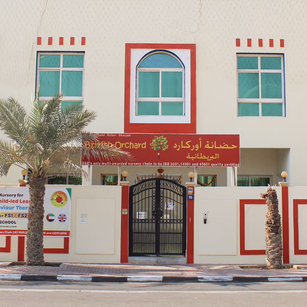 Front view of a beige and red building with three green-tinted windows, a gated entrance, palm trees, and a sign for British Orchard nursery.