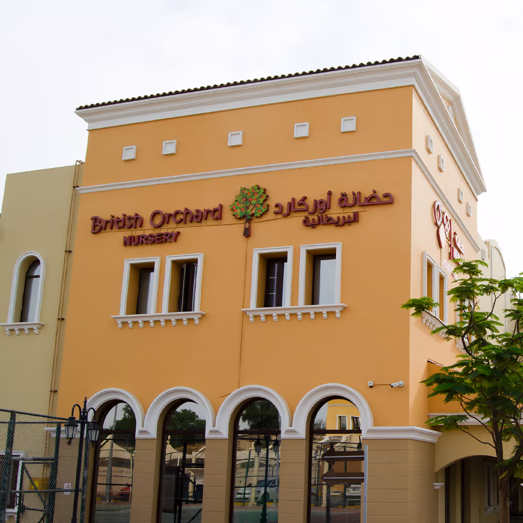Yellow building with arched windows housing British Orchard Nursery with its logo of a tree and bilingual signage in English and Arabic.