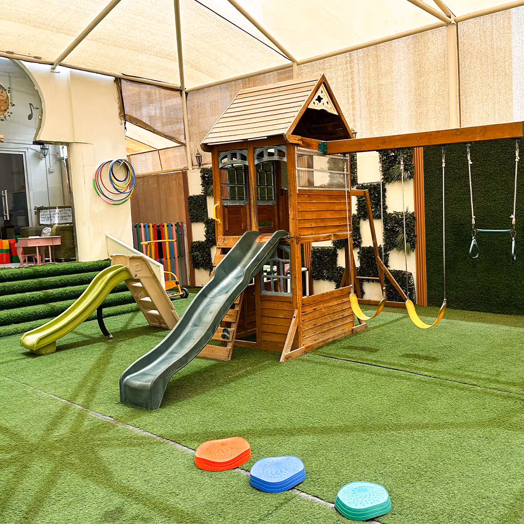 Indoor children's playground with artificial grass flooring, featuring two slides, swings, and a wooden playhouse.