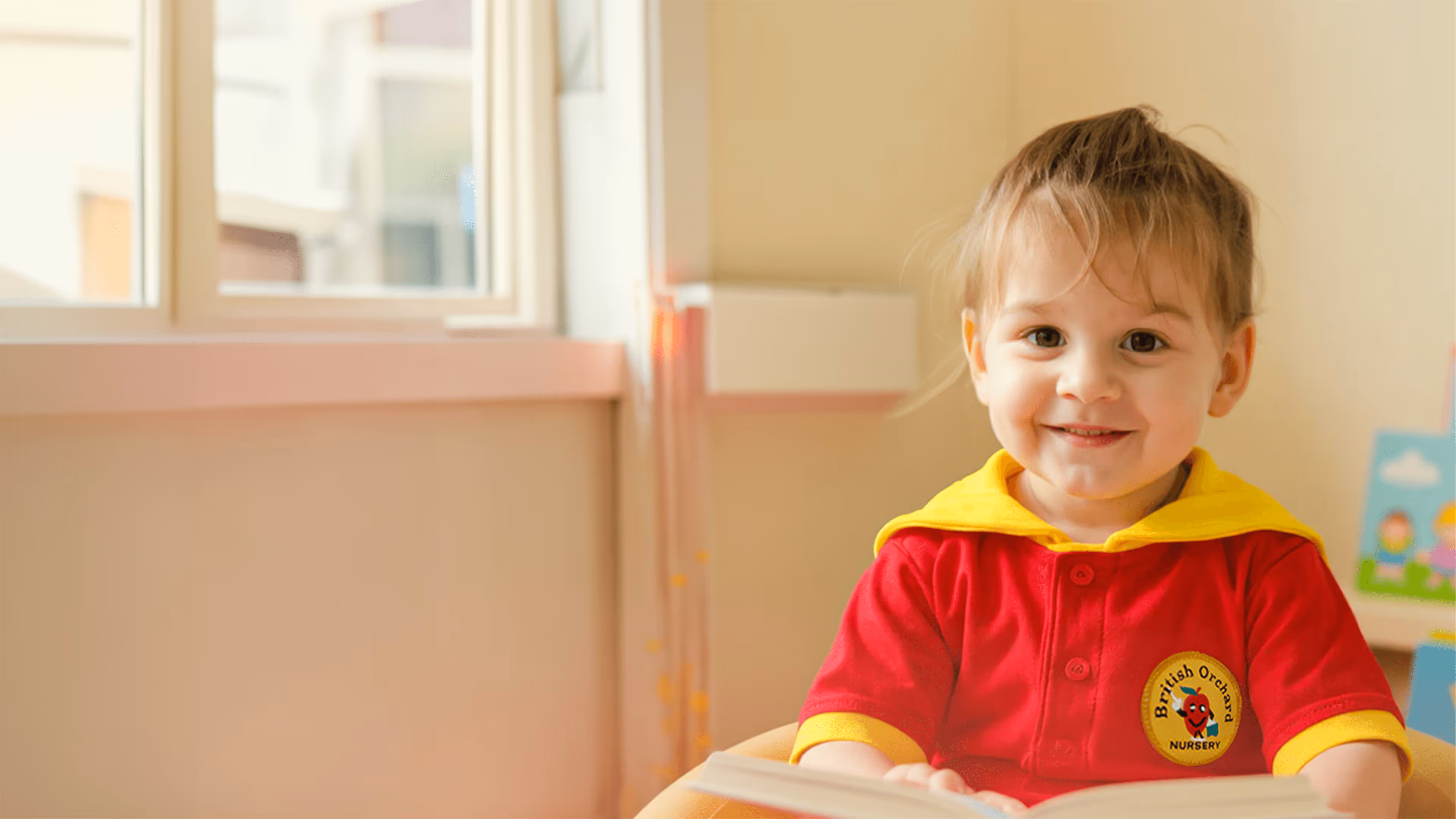 Smiling toddler wearing a red and yellow British Orchard Nursery uniform, sitting and holding an open book indoors.