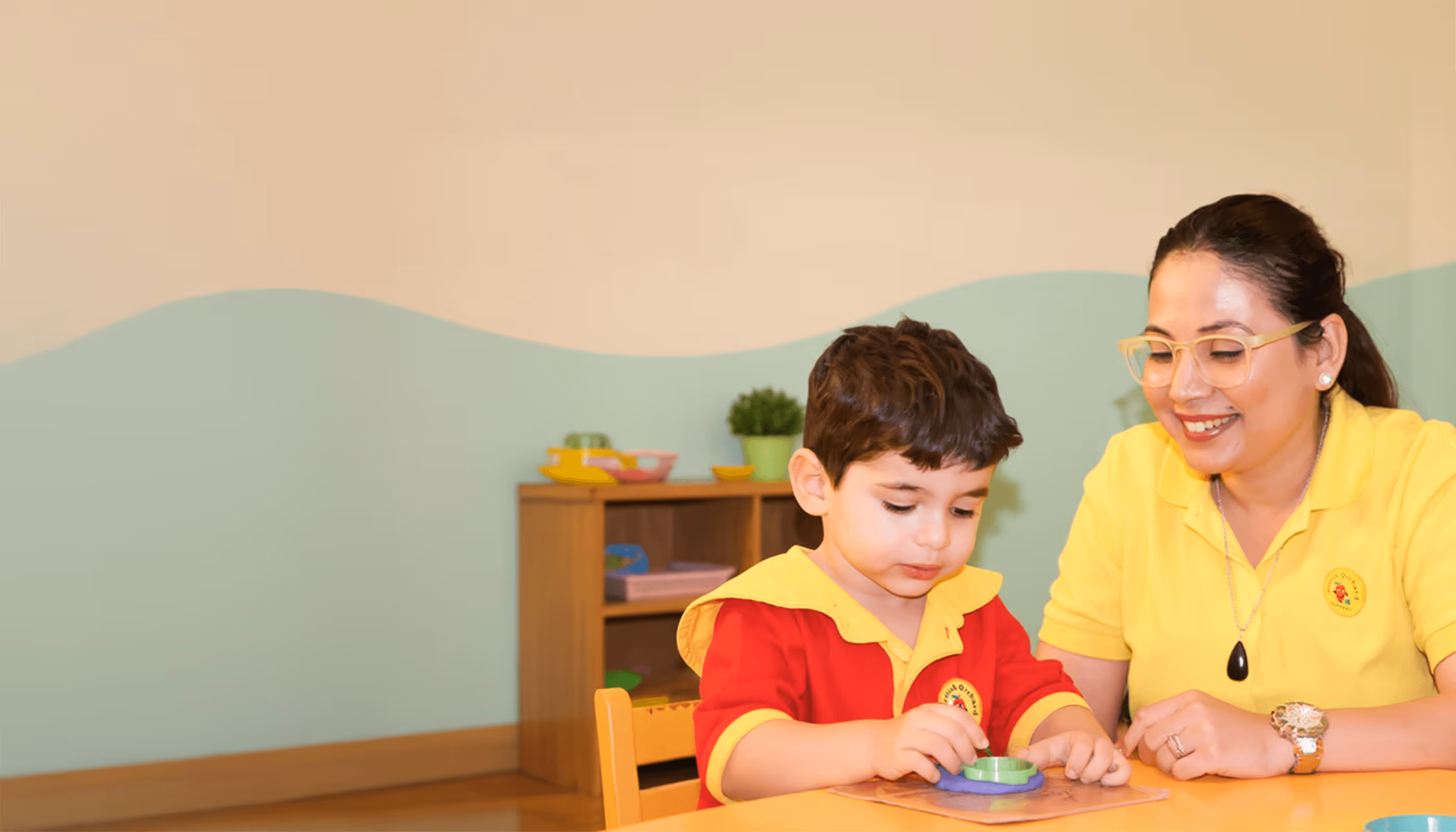 Young boy playing with a green shape sorter while a smiling teacher watches him at a table in a classroom.
