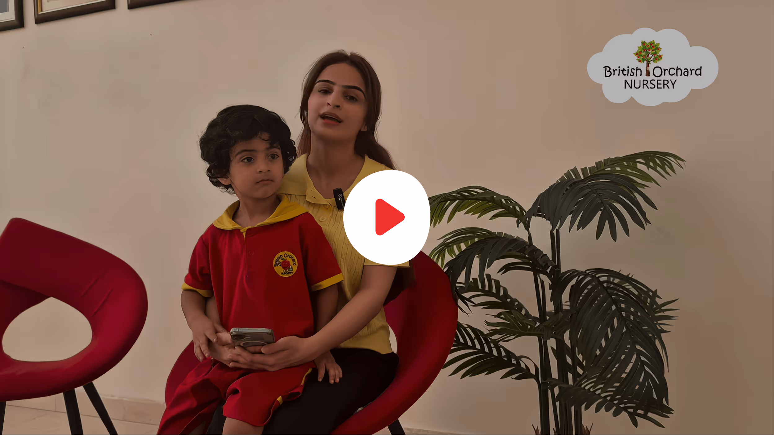 A woman in a yellow shirt sits on a red chair holding a child in a red British Orchard Nursery uniform with a smartphone, next to a green potted plant.