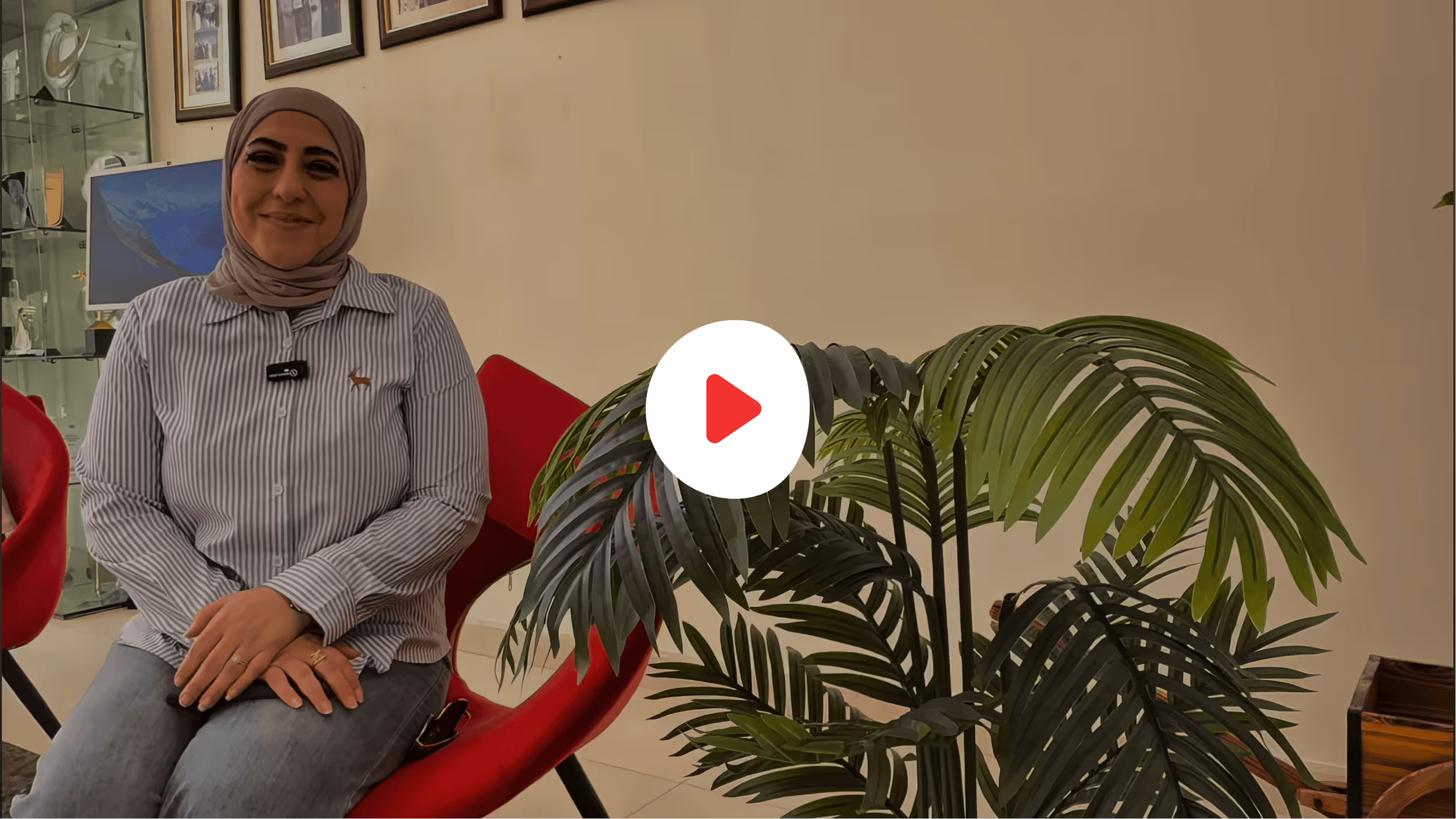 Smiling woman in a hijab and striped shirt sitting on a red chair next to a large green plant indoors.