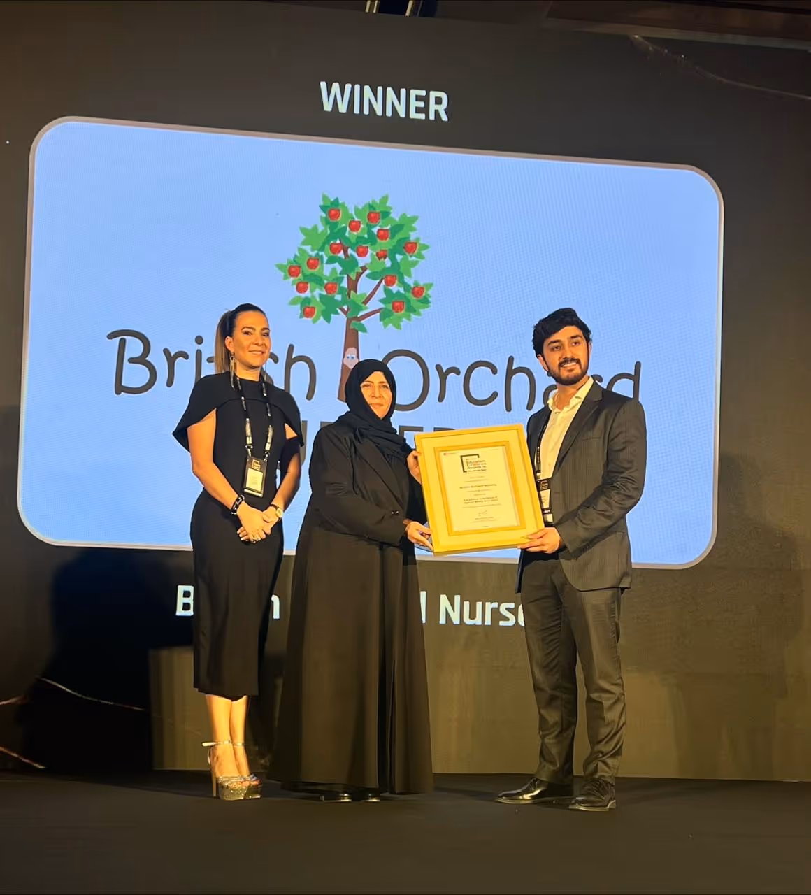 Three people on stage holding an award certificate with a large screen behind displaying the British Orchard Nursery logo and the word 'WINNER'.