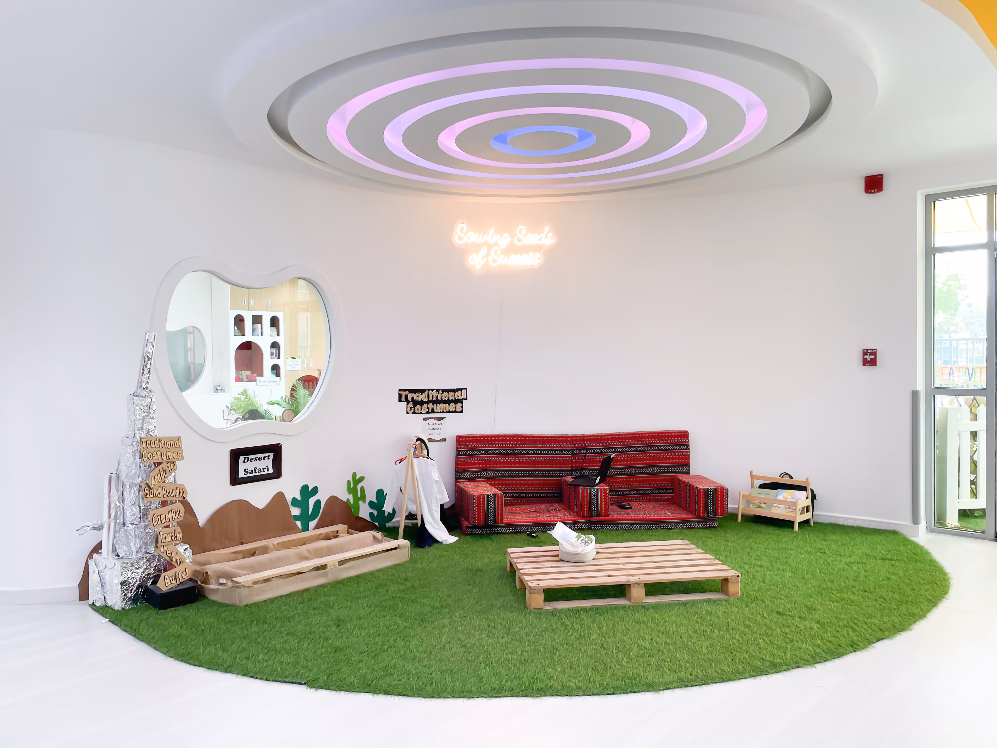 Bright indoor play area with green artificial grass, red patterned sofa, wooden pallet table, and wall signs reading 'Traditional Costumes' and 'Desert Safari'.