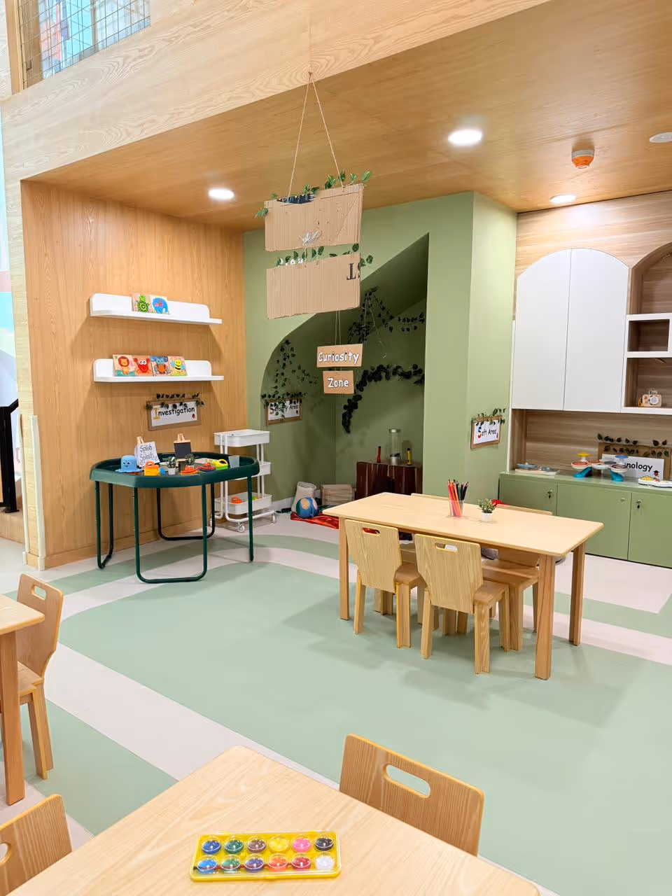 Bright children's classroom with light wood furniture, activity tables, play zones labeled Curiosity Zone, Investigation, and Soft Area, and shelves with toys and books.