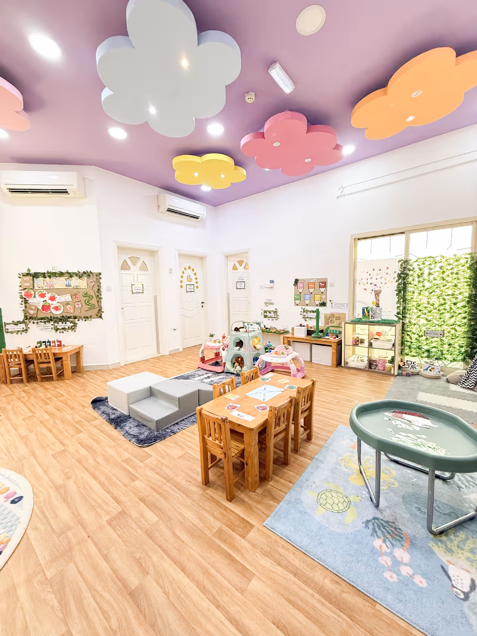 Bright and colorful kindergarten classroom with small wooden tables and chairs, playful toys, and ceiling decorations shaped like clouds and flowers.