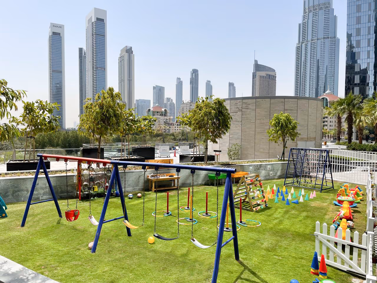 Outdoor playground with swings, climbing equipment, and colorful cones on green grass with city skyscrapers in the background under a clear sky.