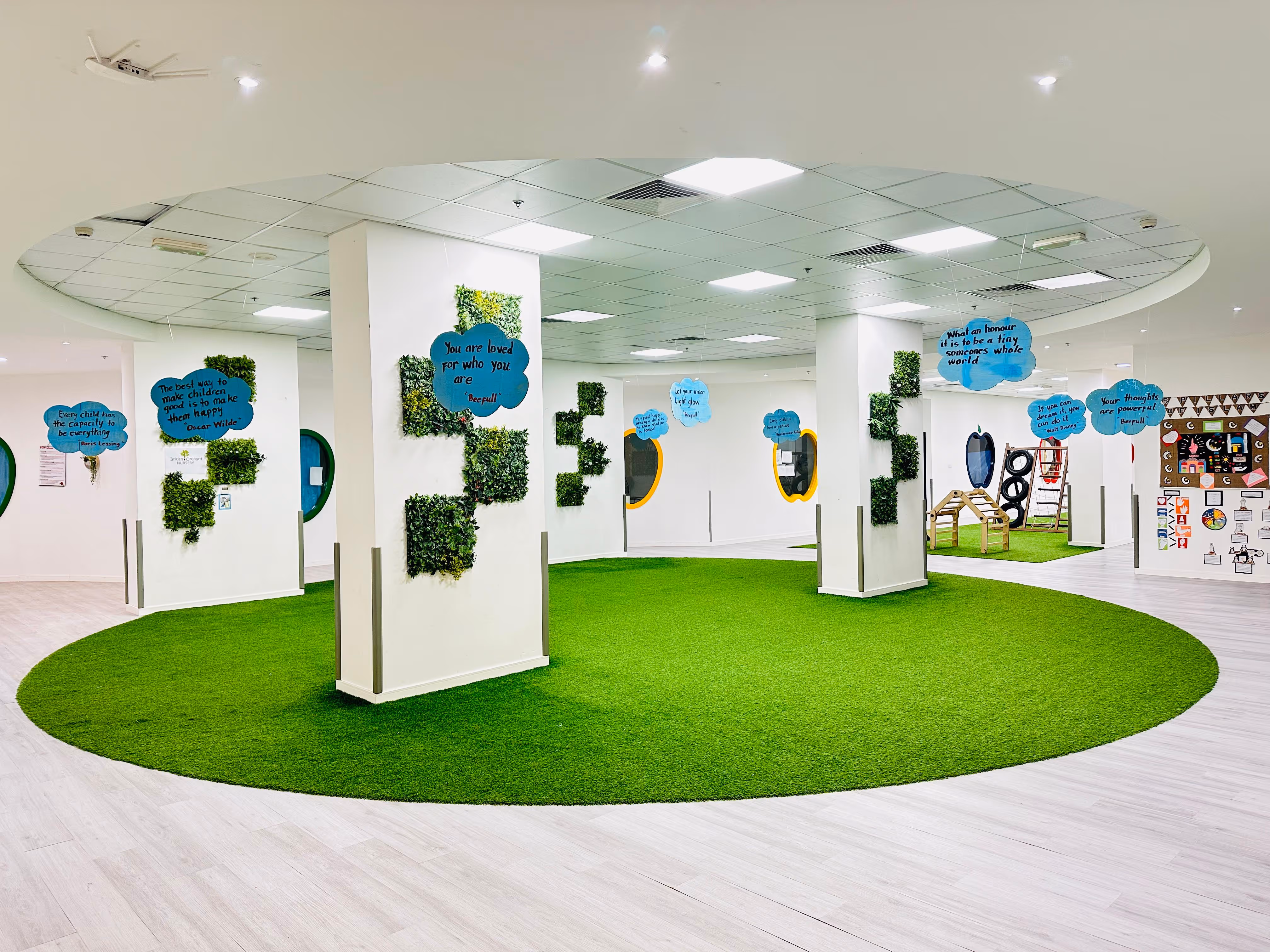 Bright indoor play area with green circular carpet, white pillars decorated with green plants, and hanging blue cloud-shaped signs featuring inspirational quotes.