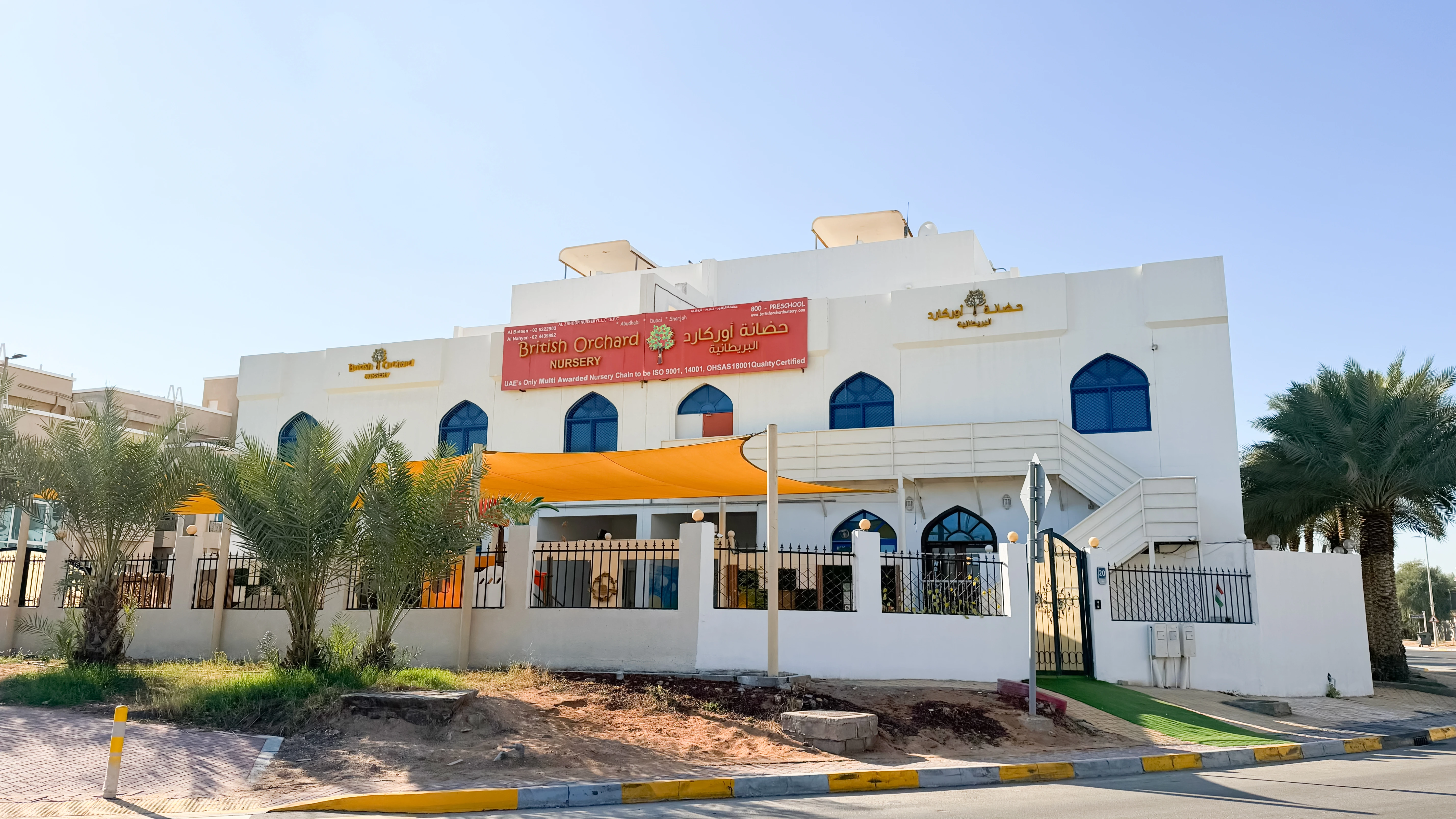 White two-story building with blue arched windows and a red banner reading British Orchard Nursery, shaded by orange canopy, surrounded by palm trees and a black iron fence.