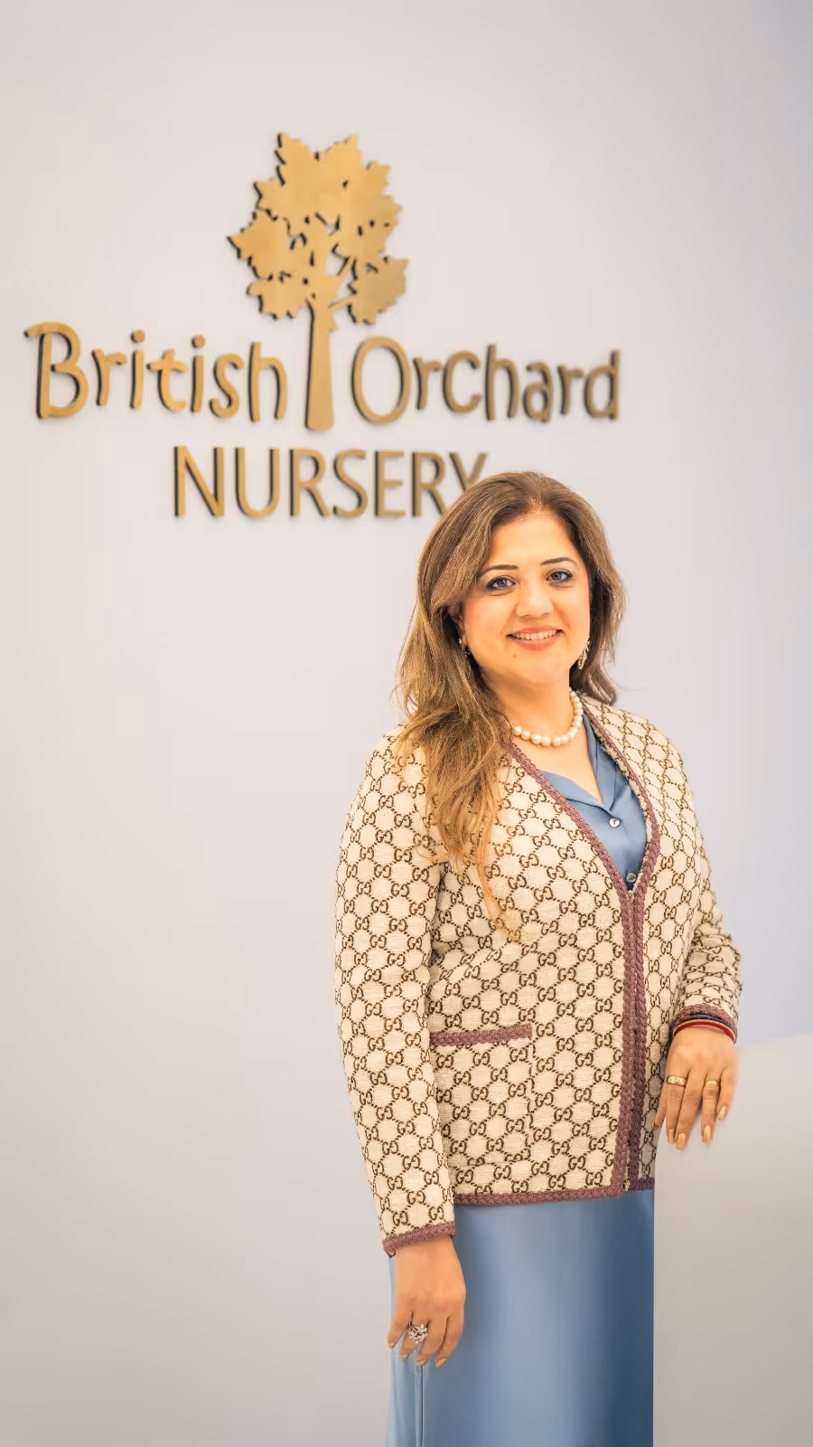 Smiling woman with long hair wearing a patterned cardigan and blue dress standing in front of a British Orchard Nursery sign.