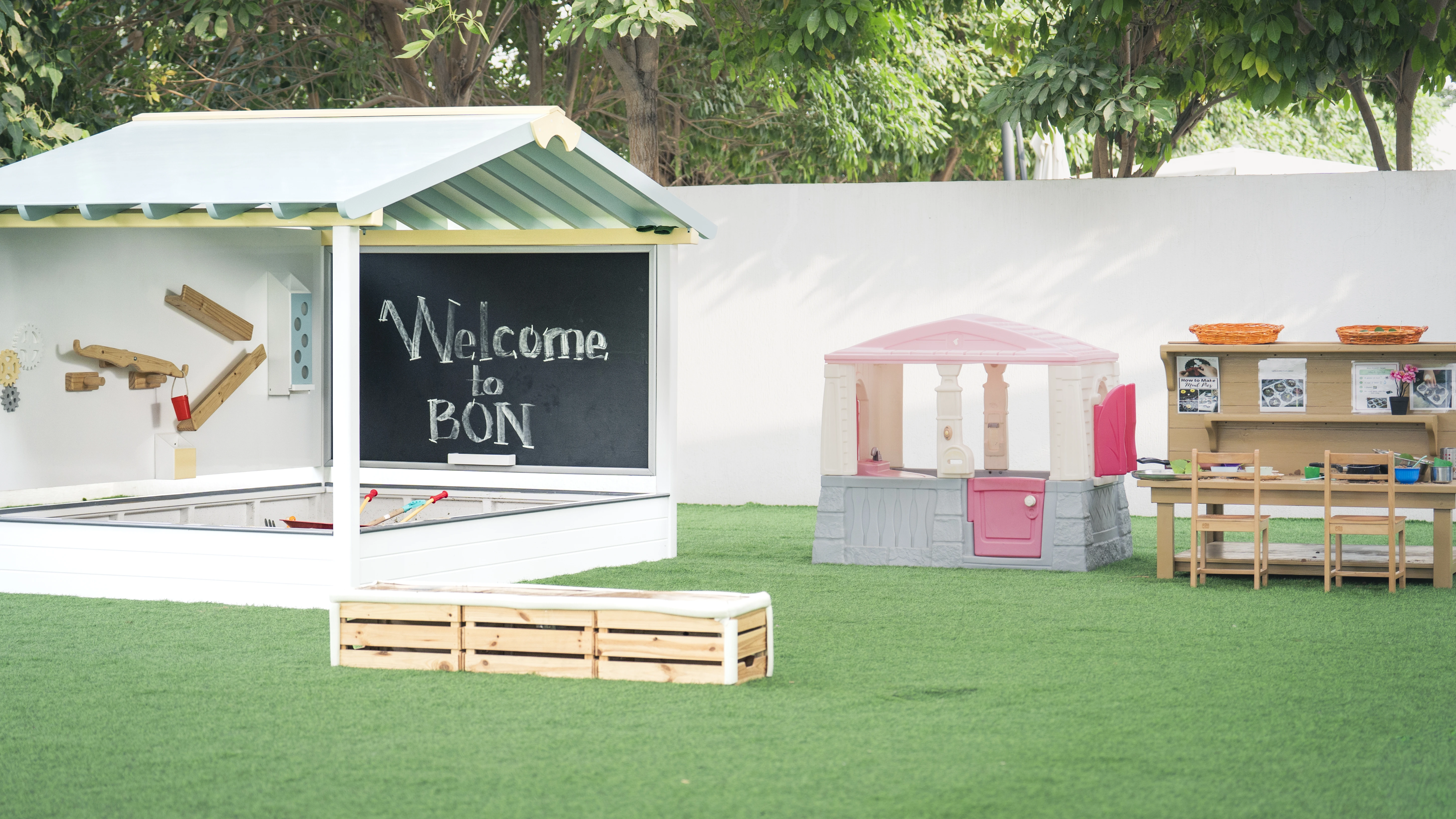 Children's outdoor play area with a chalkboard reading 'Welcome to BŌN', a small playhouse, and a wooden table with chairs and baskets.