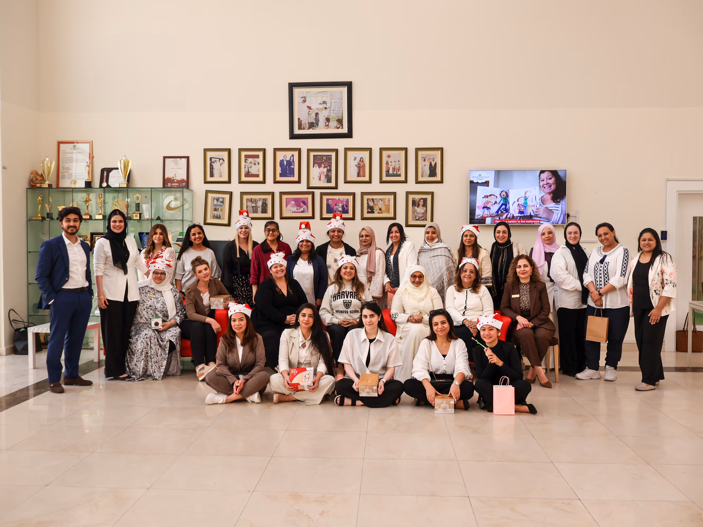 Group of diverse adults posing indoors, some wearing Santa hats, with framed photos and trophies displayed behind them.