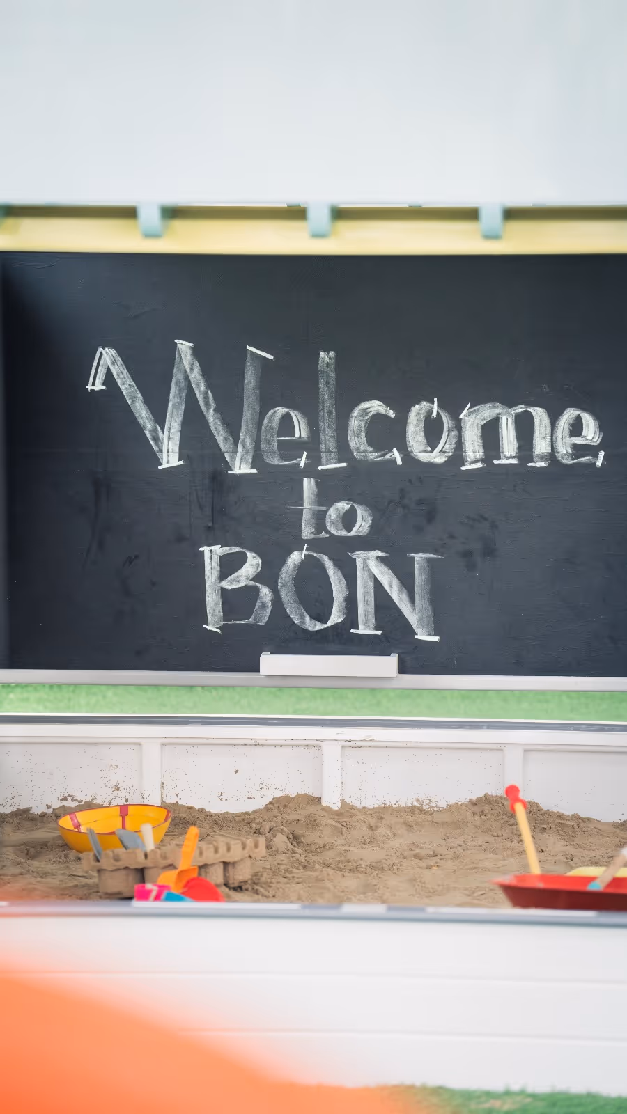 Blackboard with 'Welcome to BON' written in chalk above a sandbox filled with sand and children's sand toys.