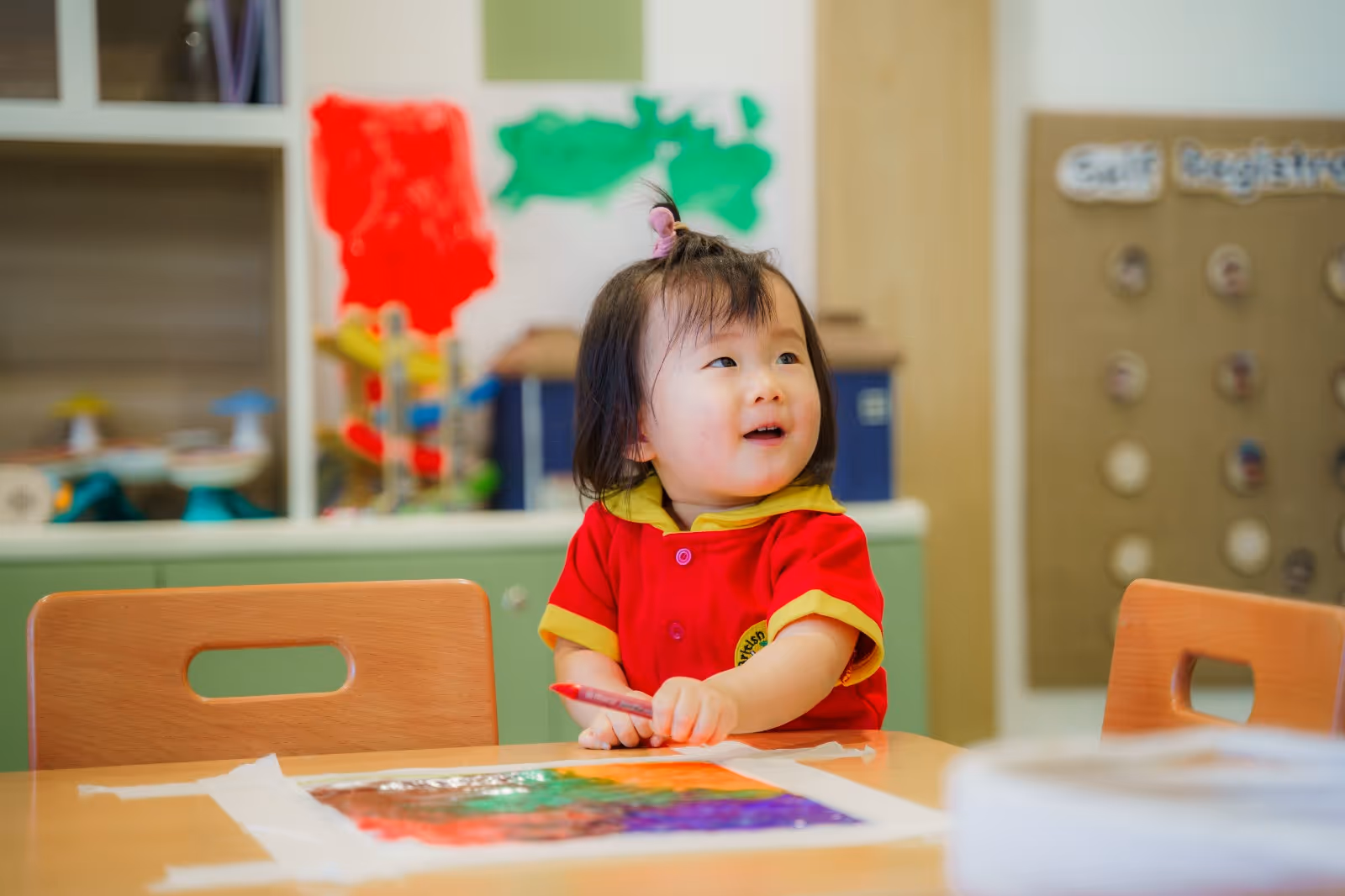 Young child in a red and yellow shirt holding a red marker, sitting at a table with colorful artwork in a classroom.